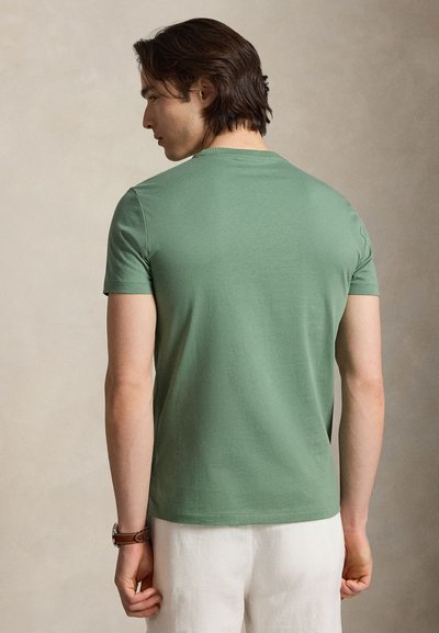 Moški s svetlo rjavimi lasmi, ki nosi zeleno kratkoročno majico in bele hlače, viden od zadaj na bež ozadju.