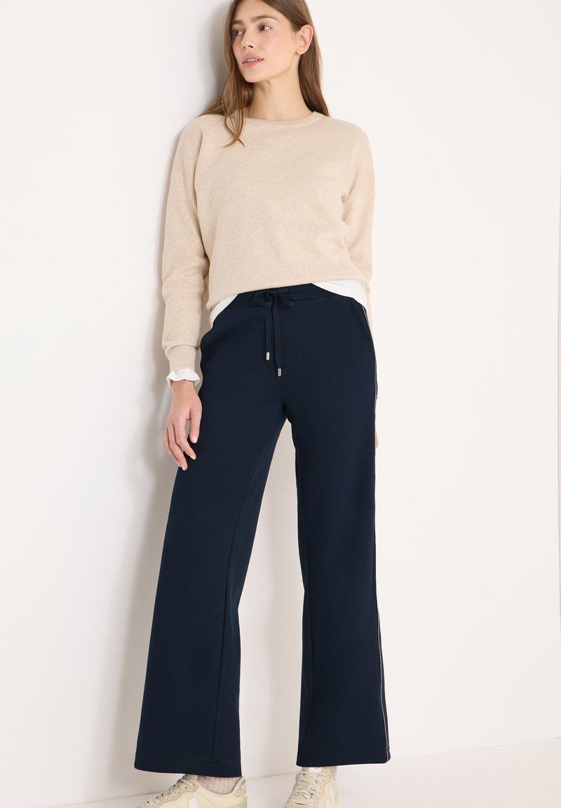 Beżowy sweter z długimi rękawami zestawiony z granatowymi spodniami o szerokich nogawkach, posiadającymi sznurek w talii i boczne kieszenie. Spodnie mają kontrastujące białe akcenty.