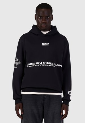 Man wearing black hoodie with text "United by a shared calling to walk the path of life bravely and boldly" and black jeans standing against a gray background.