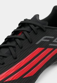 Μαύρο αθλητικό παπούτσι με υφή στην επάνω επιφάνεια, διαθέτει κόκκινες πλαϊνές ρίγες, επίπεδα κορδόνια και κομψό σχέδιο. Λαστιχένια σόλα με γωνιώδη διακοσμητικά στοιχεία.