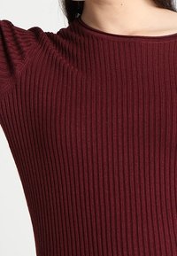 Top in tessuto a coste bordeaux con maniche corte, caratterizzato da un design aderente e una finitura liscia e testurizzata.