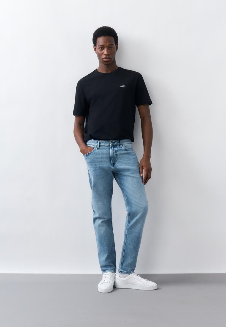 Homme portant un t-shirt noir BOSS, un jean bleu clair et des baskets blanches, debout contre un mur blanc uni avec une main dans la poche.