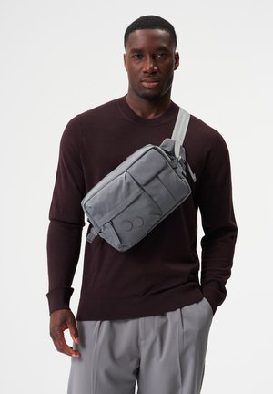 Mann trägt einen dunkel kastanienbraunen Pullover und hellgraue Hose mit einer grauen Umhängetasche über der Schulter vor einfachem Hintergrund.