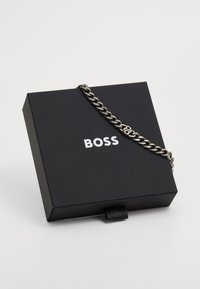 Sort gaveæske med "BOSS" i hvid tekst. Et sølvfarvet kædearmbånd ligger over boksen, hvilket viser et vævet linksdesign.