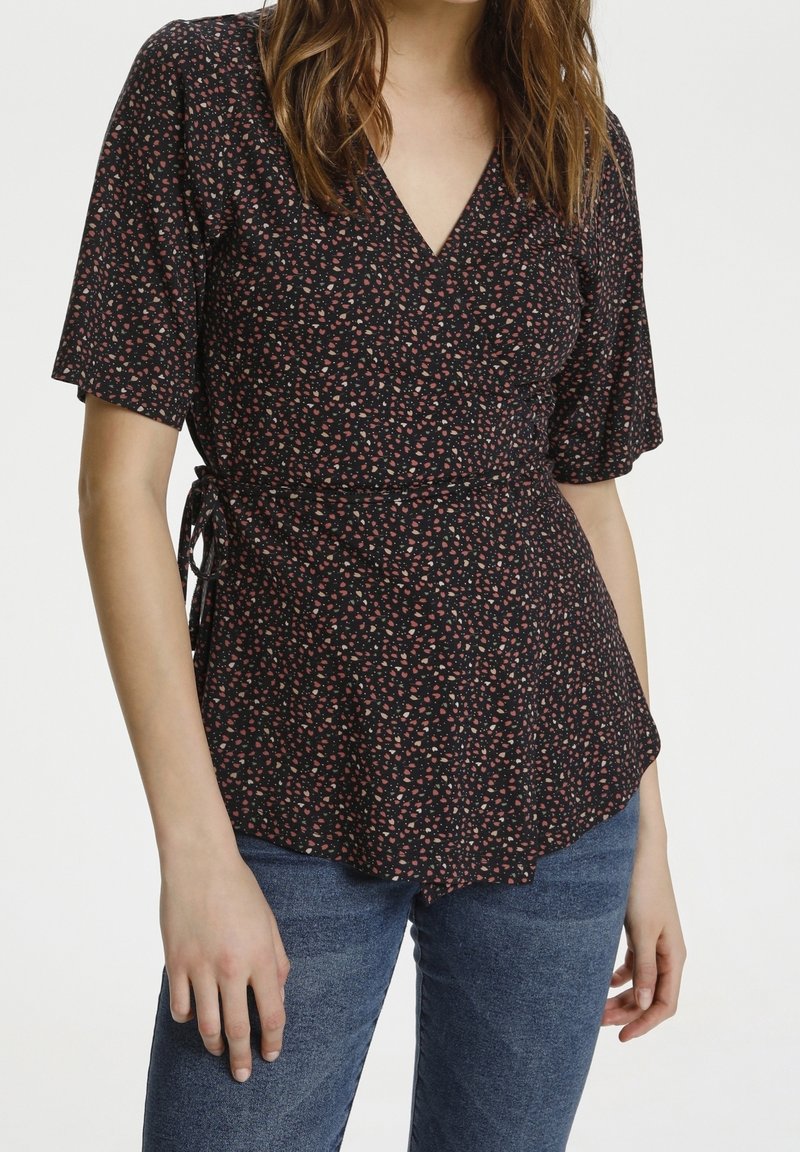 Femme portant un chemisier noir à manches courtes avec un motif floral rose et blanc et un jean bleu, debout devant un fond blanc uni.