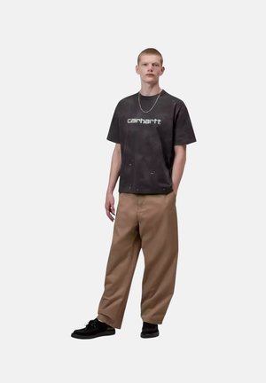 Joven con camiseta negra con logo de Carhartt, pantalones sueltos color beige, zapatillas negras y un collar de cadena plateado, de pie frente a un fondo blanco.