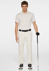 Beige pikétröja med tre knappar, svarta detaljer på axlarna, parat med vita byxor och svart bälte. Bär svarta golfhandskar och sneakers.