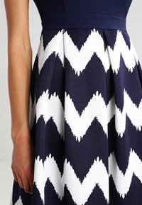 Marinen klänning i navy med ett vitt zickzackmönster, tillverkad av texturerad tyg med veck, som framhäver en figursydd bodice och en flödande kjol.