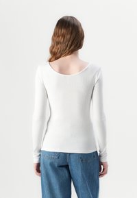 Biała, długa koszulka z prążkowanej tkaniny z szerokim dekoltem, zestawiona z jasnoniebieskimi jeansami z wysokim stanem. Widok z tyłu ukazuje dopasowany krój i teksturowany materiał.