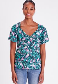 Blumenbluse in Grün mit rosa und weißen Mustern. Mit Puffärmeln und V-Ausschnitt. Getragen mit hellblauen Jeans. Glatte Textur.