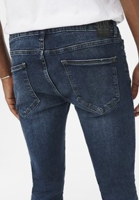 Sötétkék slim fit farmernadrág, két hátsó zsebbel és sárga varrással. A derékpántján márkázott címke található.