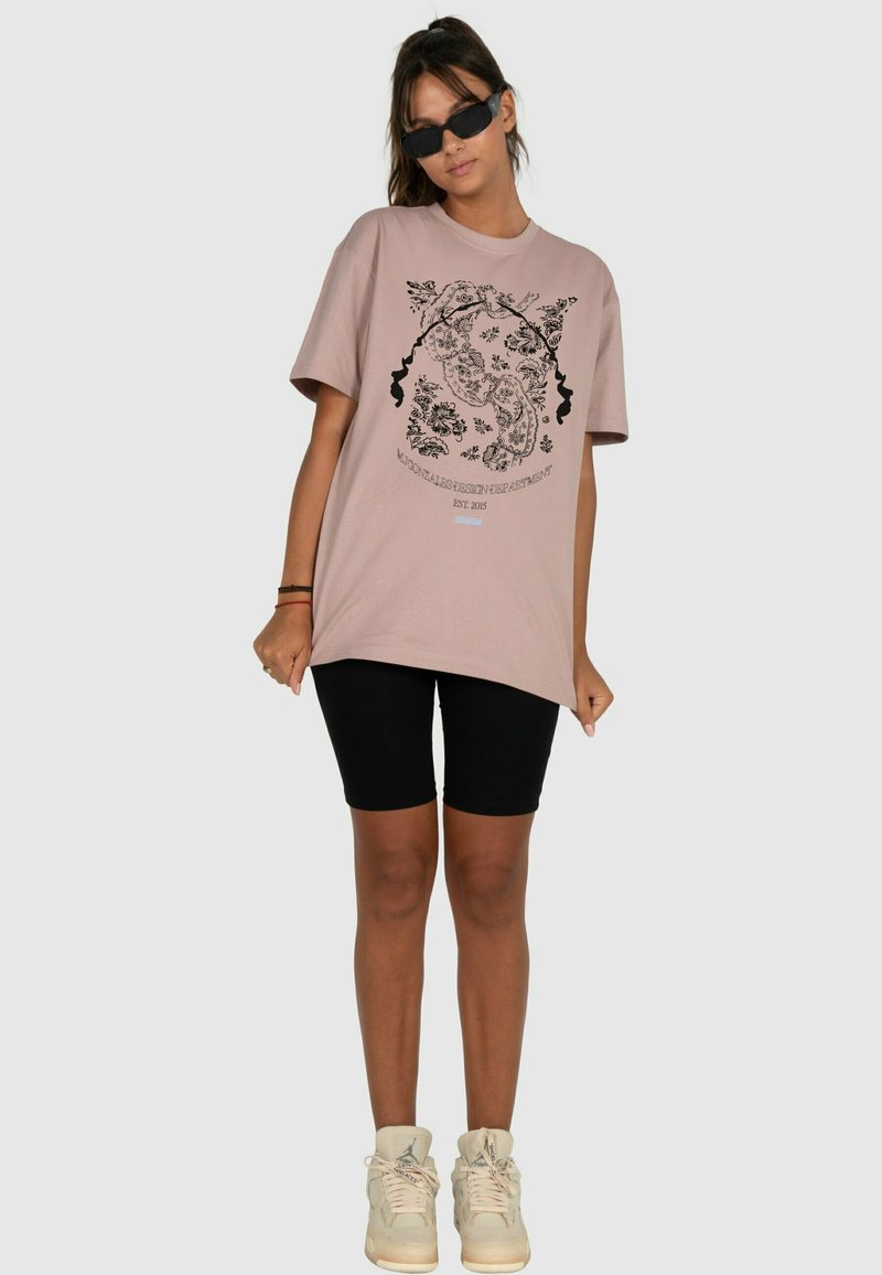 Růžové oversized tričko s černou květinovou a obličejovou grafikou. Spárováno s černými cyklistickými šortkami a béžovými teniskami.