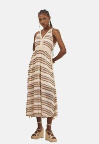 Vestido maxi en beige con patrones geométricos en marrón. Presenta un escote halter, cintura ajustada y falda acampanada. Adornado con borlas.