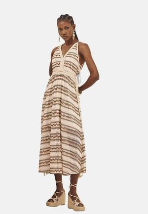 Vestido maxi en beige con patrones geométricos en marrón. Presenta un escote halter, cintura ajustada y falda acampanada. Adornado con borlas.
