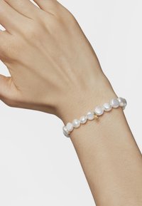 Pulsera de perlas blancas con perlas de diferentes tamaños y formas, acentuada por un pequeño charm de oro, llevada en una muñeca de piel clara.