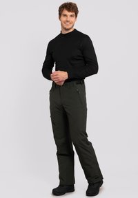 Camisa negra de manga larga y pantalones de nieve verdes oscuros con cintura ajustable, bolsillos con cremallera y un tejido texturizado. Calzado negro completa el atuendo.