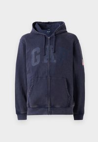 Felpa con zip di colore blu navy, con tasche frontali, logo GAP sul petto, cappuccio con coulisse e una piccola toppa della bandiera americana sulla manica sinistra.