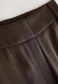 Gros plan sur un pantalon en tissu marron foncé avec une ceinture lisse munie d'un élastique froncé à l'arrière et un détail de couture centrale.