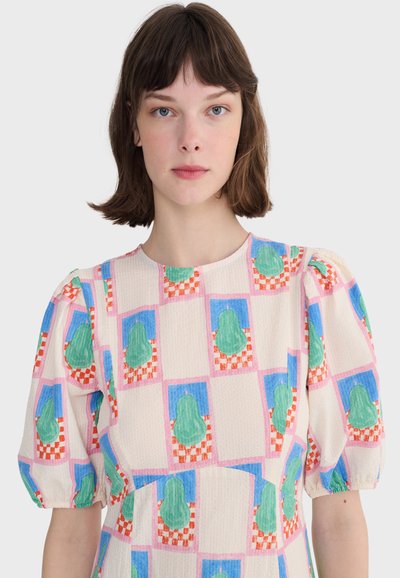 Femme aux cheveux courts bruns portant une robe à manches bouffantes, ornée de motifs de poires vertes sur un fond en carrés bleus et roses à carreaux blancs.