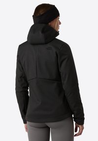 Svart huva jacka med en texturerad yta. Har en framträdande söm på baksidan och logodetalj, kompletterad av en åtsittande design och handledsmuddar.