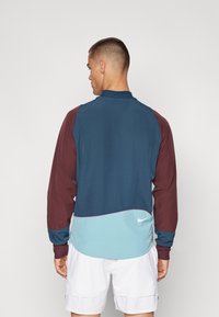 Chaqueta Nike con un diseño a bloques de colores que combina secciones en azul oscuro, burdeos y azul verdoso. De manga larga, con cuello alto y un detalle sutil del logo.