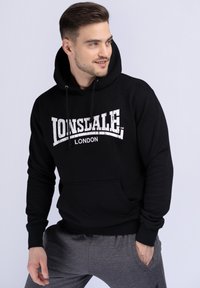 Čierna mikina s kapucňou a veľkým bielym potlačou "LONSDALE LONDON". Obsahuje predné vrecko a ribbované manžety, vyrobená z mäkkého materiálu.