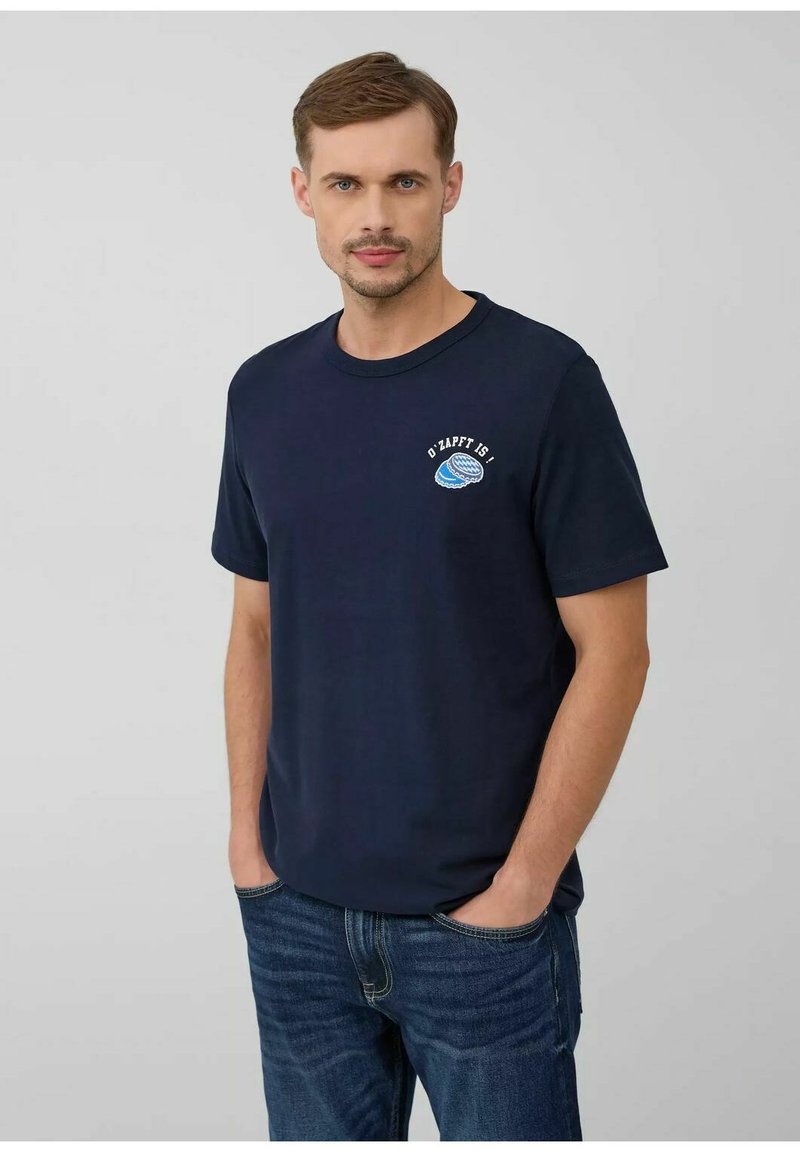 Homme aux cheveux châtain clair portant un t-shirt bleu marine avec un logo blanc et bleu sur la poitrine et un jean bleu, mains dans les poches, fond neutre.