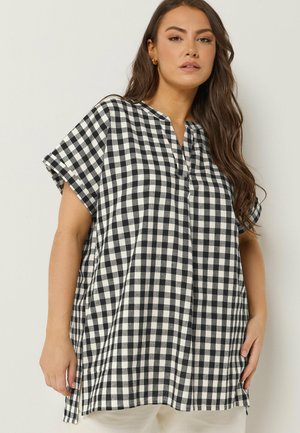 Frau trägt eine schwarz-weiß karierte Kurzarm-Tunika mit kleinem V-Ausschnitt und Seitenschlitzen, steht vor einem einfarbigen Hintergrund.