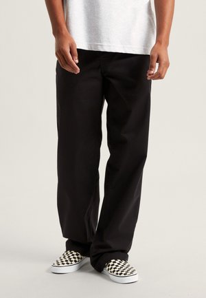 Person trägt schwarze, weit geschnittene Hose, weißes T-Shirt und schwarz-weiß karierte Slip-On-Schuhe und steht vor einem schlichten Hintergrund.