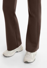 Beine in dunkelbraunen, weiten Hosen und off-white chunky Sneakers stehen vor einem schlichten weißen Hintergrund.