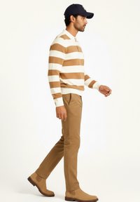 Suéter a rayas en beige y blanco, pantalones beige y botas de tobillo beige. Modelo lleva una gorra azul marino, caminando de perfil. Tejido texturizado.