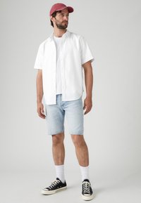Cămașă albă cu mânecă scurtă, pantaloni scurți din denim bleumarin, adidași negri cu talpă albă și o șapcă roșie. Ținută de vară casual.
