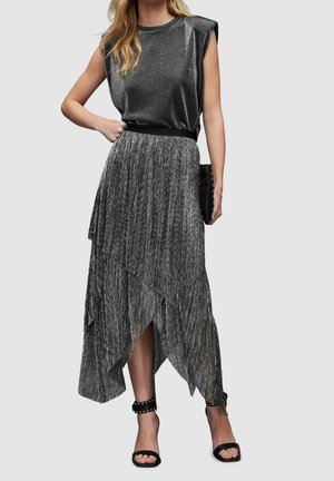 Žena v bezrukávovém třpitivém černém topu a asymetrické metalické plisované sukni, s černými kotníkovými podpatky s nýtky, nesoucí černou kabelku do ruky.