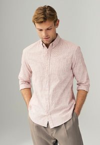 Lys pink skjorte med knapper og et subtilt stribemønster, korte ærmer rullet op, og en enkelt brystlomme, parret med lysegrå bukser.