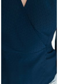 Navyblaues Wickeloberteil aus strukturiertem Stoff, mit tiefem V-Ausschnitt und diagonaler Naht. Das Material erscheint weich und glatt.