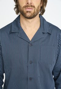 Camisa de pijama de rayas azul marino con cuello corto, cierre de botones y un solo bolsillo en el pecho. Fabricada en un tejido suave y ligero con rayas verticales.
