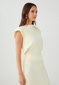 Top senza maniche di un delicato giallo chiaro con scollatura alta, vita aderente e texture a coste, abbinato a una gonnellina plissettata coordinata.