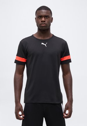 Νέος άνδρας με μαύρο μπλουζάκι Puma με κόκκινες ρίγες στα μανίκια, στέκεται με το σώμα προς τα εμπρός μπροστά σε απλό λευκό φόντο.
