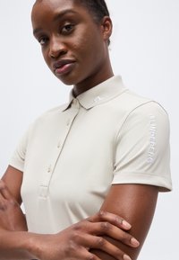 Γυναίκα με σταυρωμένα χέρια, φοράει μπεζ πόλο μπλούζα J.Lindeberg με λογότυπο στο γιακά και το μανίκι, μπροστά σε απλό φόντο.