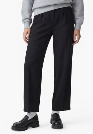 Persona con pantalones negros de pierna recta, sudadera gris, calcetines blancos y mocasines gruesos negros, de pie delante de un fondo blanco.