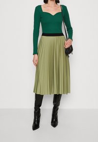 Top de manga larga verde acanalado con escote cuadrado, combinado con una falda plisada verde oliva. Terminado con botas negras hasta la rodilla y un pequeño bolso negro.