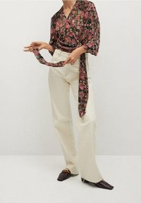 Kvinde iført crèmerfarvede wide-leg jeans, blomsterwrap-bluse med bindebånd og spidse, mørkebrune sko med gyldne kæder.