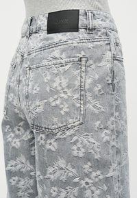 Κοντινό πλάνο ανοιχτόχρωμων γκρίζων τζιν με ανάγλυφη κεντητή λουλουδάτη διακόσμηση και μαύρη δερμάτινη ετικέτα επωνυμίας στη μέση.