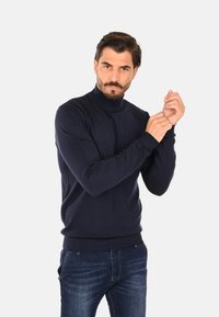 Maglione dolcevita blu navy in tessuto morbido, con maniche lunghe e polsini a coste. Abbinato a jeans in denim scuro.