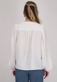 Witte, gestructureerde blouse met pofmouwen en een subtiele glans; achterkant heeft een yoke en plooien langs de halslijn. Gecombineerd met blauwe broeken.
