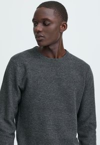 Grå strikket genser med rund hals, med en teksturert overflate og ribbestrikkede mansjetter. Stoffet ser mykt og middels tungt ut.