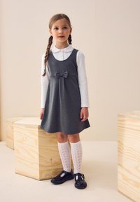 Abito grigio senza maniche con dettaglio a fiocco, abbinato a una camicia bianca a maniche lunghe e calze bianche a motivi alte fino al ginocchio. Scarpe nere.