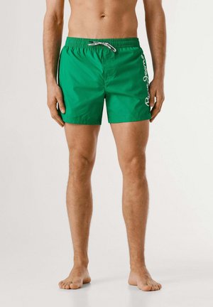 Uomo in piedi a piedi nudi che indossa pantaloncini da bagno verdi con coulisse bianca e logo laterale su sfondo semplice.