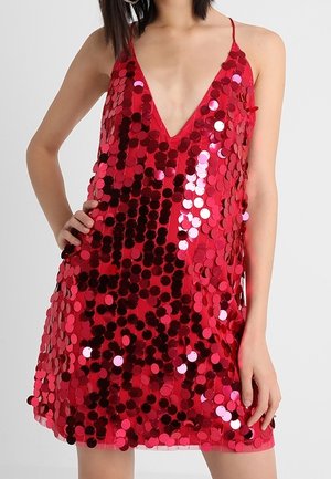 Vrouw met een mouwloze rode mini-jurk bedekt met grote glanzende pailletten, poseert met één hand op de heup en de andere ontspannen.