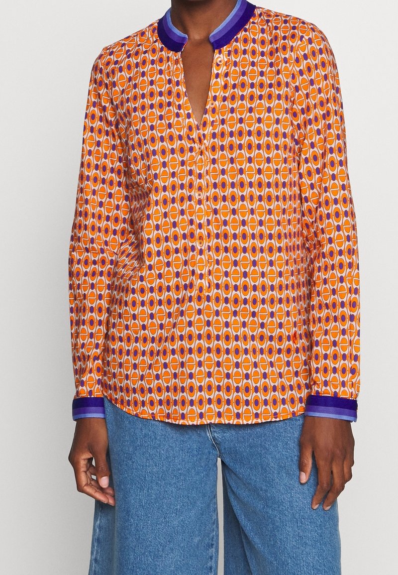 Personne portant un chemisier orange à manches longues avec un motif géométrique violet et blanc, col et poignets violets, associé à un jean en denim bleu.